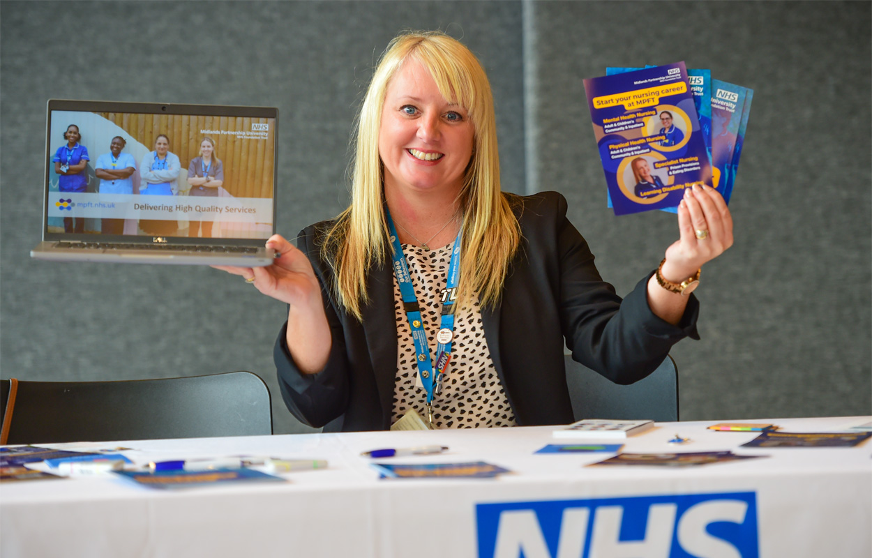 A smartly dressed female NHS employee is sat at a table with information about job opportunities