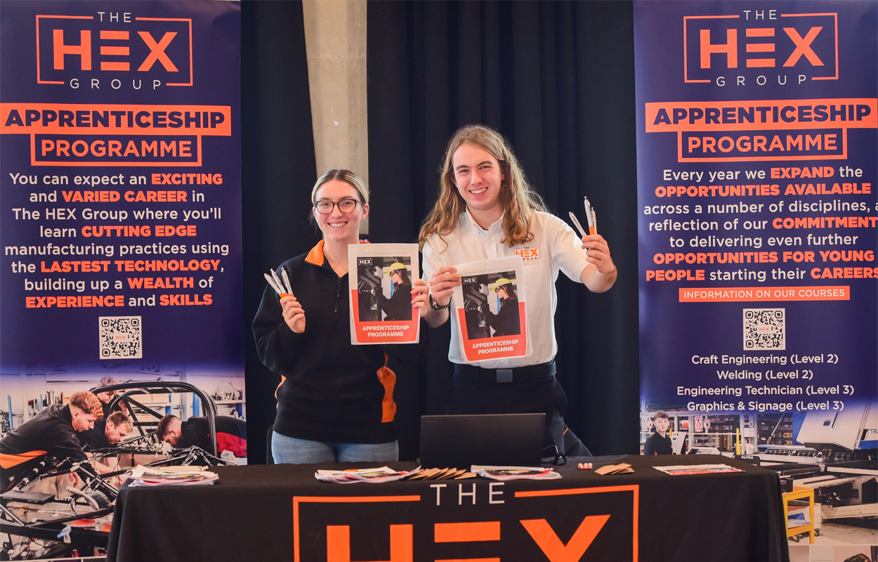 Female and male employees dressed in casual uniforms are stood at an information stand