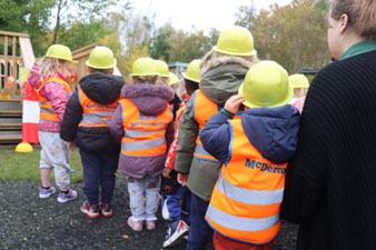 Nursery children are wearing hi-vis jackets and helmets outside on the playground