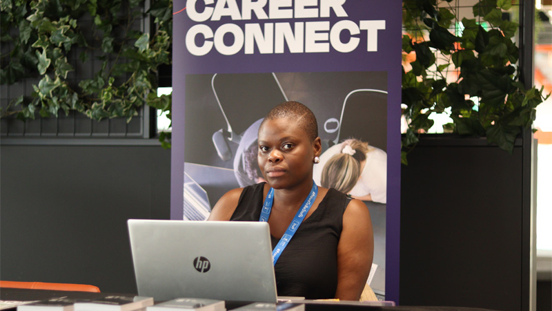 A female wearing dark clothing is sitting with a laptop in front of a careers stand