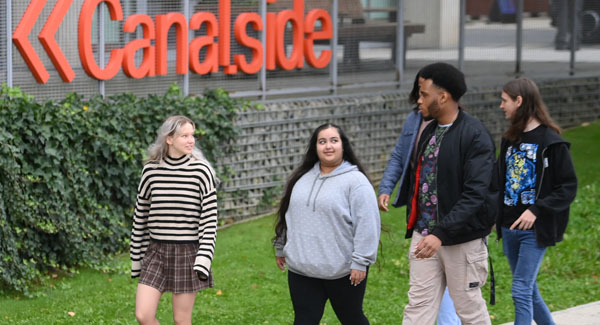 A group of young students dressed casually are talking and walking towards Canalside near the London Campus