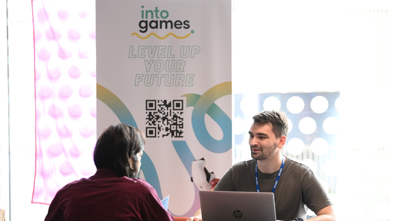 Two males dressed casually are chatting in front of an Into Games stand