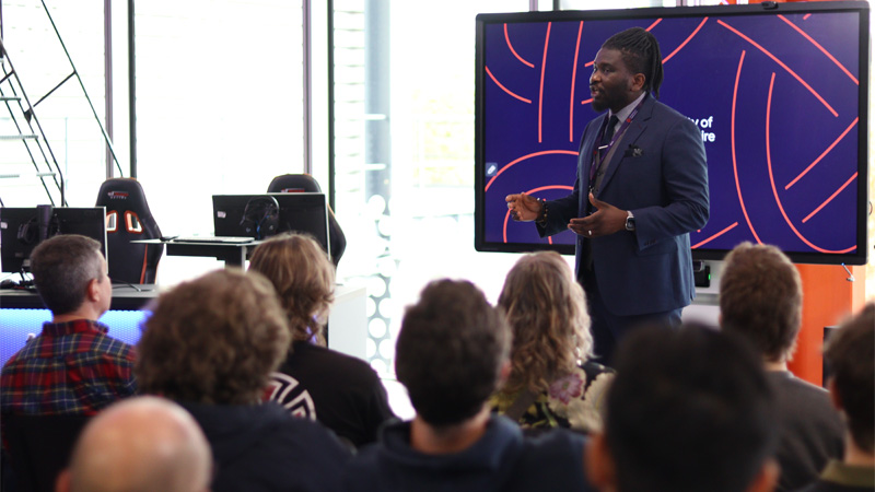 A senior staff member dressed formally is stood up delivering a talk to an audience of prospective students and parents