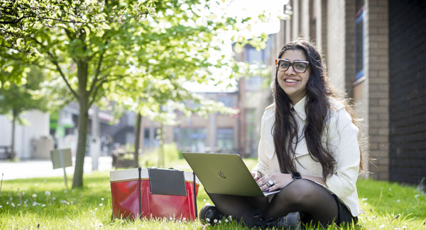 A female student with a laptop is sitting on the grass smiling