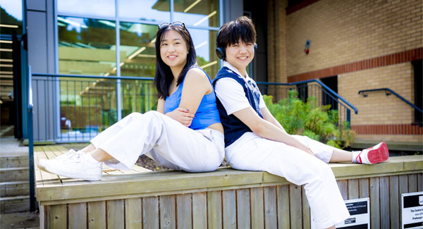 A female and male student dressing casually are sitting on a wooden bench posing for a photo outside the Stafford Centre building