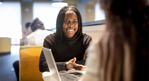 A young female with long black hair is sitting at a table with a laptop chatting to another female